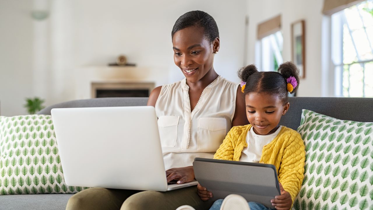 Woman sitting on sofa working on computer with daughter by her side.